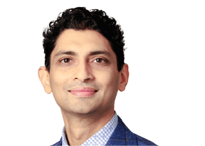 A person with short curly hair wearing a blue checkered shirt and a blue blazer is smiling against a white background. Description generated by AI