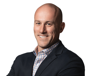 A smiling adult with a shaved head wearing a dark blazer over a checkered shirt stands with arms crossed against a white background. Description generated by AI